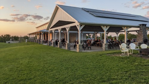 A spacious outdoor venue with a covered patio, people dining, and a sunset in the background over green grass.