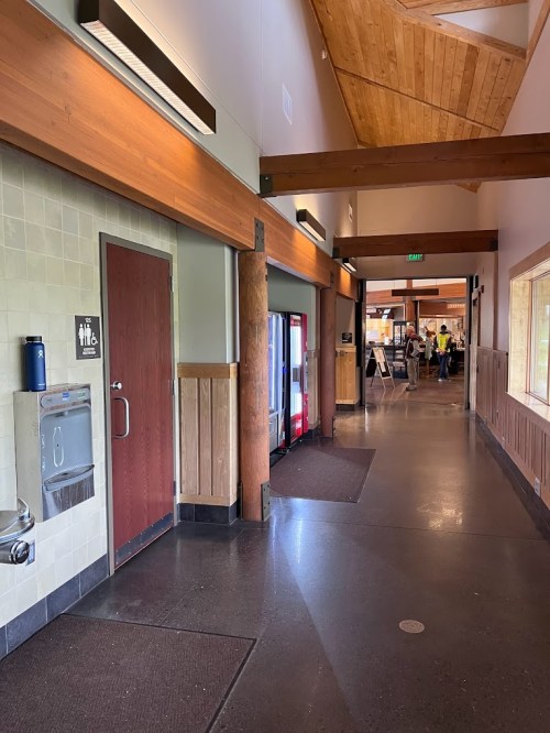 Interior hallway with wooden beams, restrooms on the left, a water fountain, and a glimpse of a kitchen area ahead.