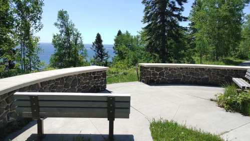 A scenic view of a lake from a stone bench area surrounded by trees and greenery on a sunny day.