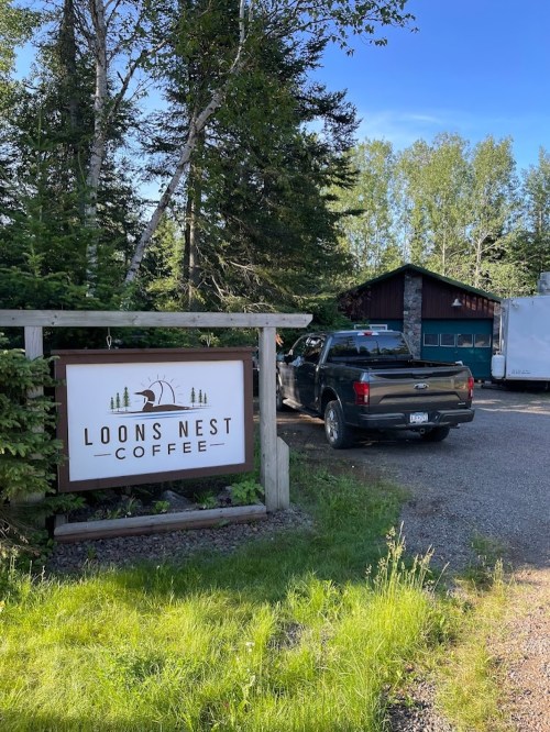 A sign for "Loons Nest Coffee" near a rustic building and parked truck, surrounded by trees and greenery.