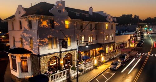 A charming hotel at dusk, with warm lights glowing and cars passing by on a quiet street.