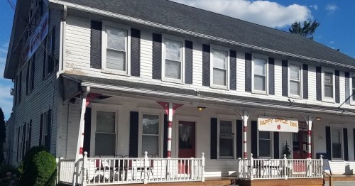 A two-story white building with black shutters and a porch, featuring a sign that reads "Happy Apple Inn."