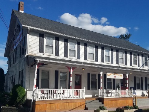 A two-story white building with black shutters, a porch, and a sign reading "Happy House 1903" under a blue sky.