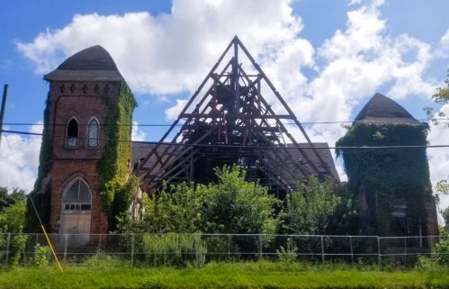 Abandoned brick building with ivy-covered towers and a partially collapsed roof under a blue sky with clouds.