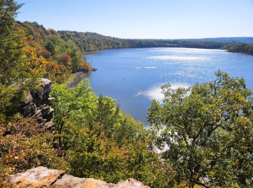 A serene lake surrounded by colorful autumn trees under a clear blue sky. Sunlight sparkles on the water's surface.