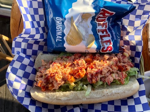 A lobster roll filled with lobster meat on a bed of lettuce, served with a bag of Ruffles potato chips.