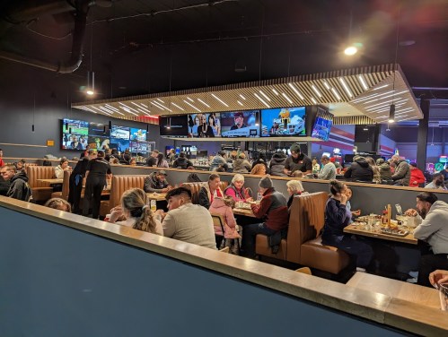 A busy restaurant with patrons seated at booths, watching screens and enjoying food in a lively atmosphere.