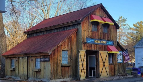 A rustic wooden building with a red roof, featuring a "Mini-Golf" sign and an American flag outside.