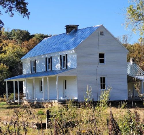 A white two-story house with a blue metal roof, surrounded by trees and greenery, set in a rural landscape.