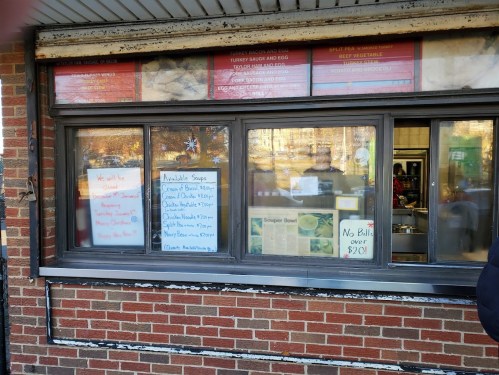 A takeout window with a menu and signs displaying daily specials and soup options, set against a brick wall.