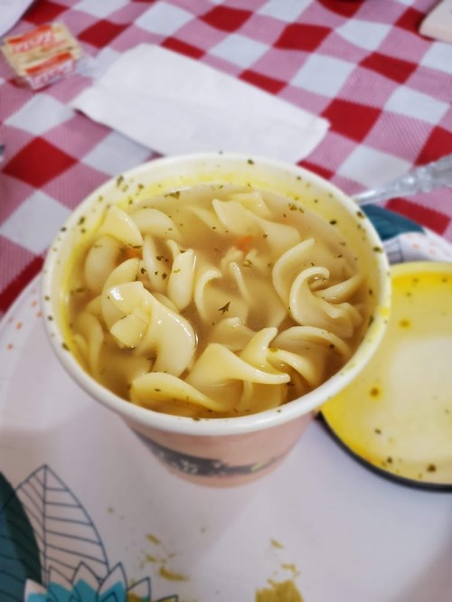 A cup of noodle soup with spiral pasta and herbs, placed on a patterned plate with a checkered tablecloth.