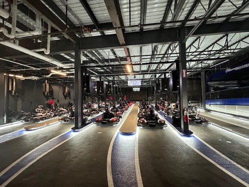 Indoor go-kart track with multiple karts lined up, illuminated by blue and white lights, under a metal roof.