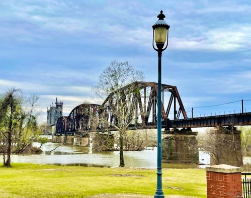 A rusty railway bridge spans a river, with a lamppost and trees in the foreground under a cloudy sky.