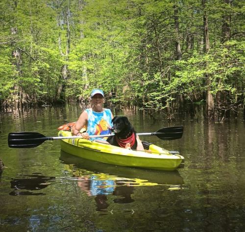 A person in a kayak with a dog, surrounded by lush green trees and calm water.