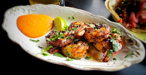 Grilled shrimp garnished with herbs, served with a lime wedge and a side of orange sauce on a decorative plate.
