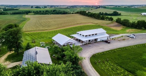 Aerial view of a modern building surrounded by green fields and vineyards under a colorful sunset sky.