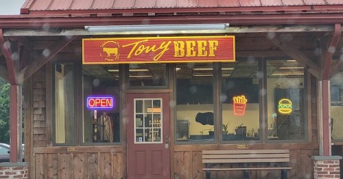 Exterior of Tony Beef restaurant with neon signs for "Open," fries, and a burger. Wooden structure with a red roof.