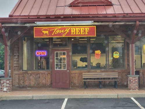 A rustic restaurant exterior with a red roof, featuring a sign that reads "Tony Beef" and neon lights for open, fries, and burgers.