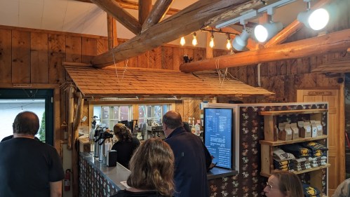 A cozy coffee shop interior with wooden beams, a counter, and customers waiting in line.