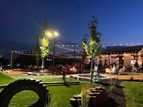 A lively outdoor space at night with string lights, people socializing, and a playground area.
