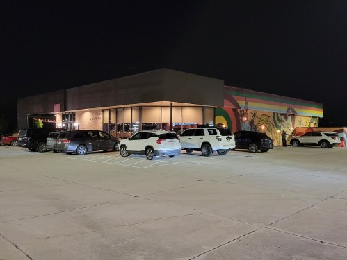 A dimly lit building at night with colorful murals, surrounded by parked cars in a spacious lot.