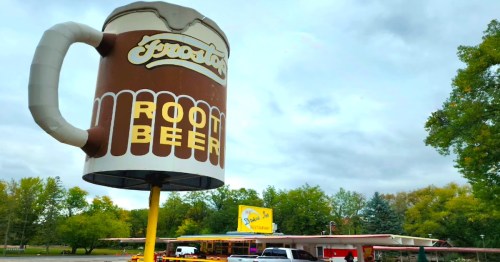 A large foam root beer mug sign in front of a retro-style restaurant with trees in the background.