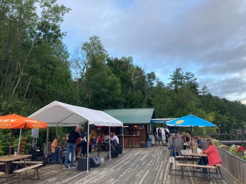 Outdoor gathering at a lakeside venue with tents, seating, and people enjoying live music amidst trees.