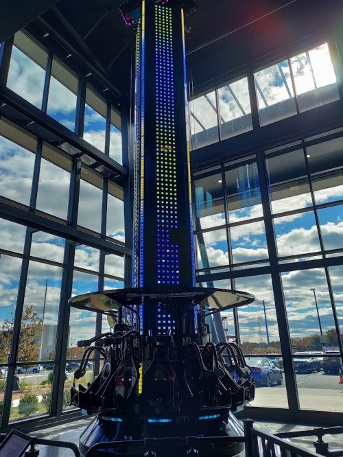 A tall amusement ride with colorful lights, set against large windows and a cloudy sky outside.