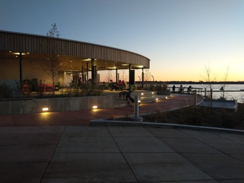 A modern building by a lake at sunset, with people sitting outside and trees lining the walkway.