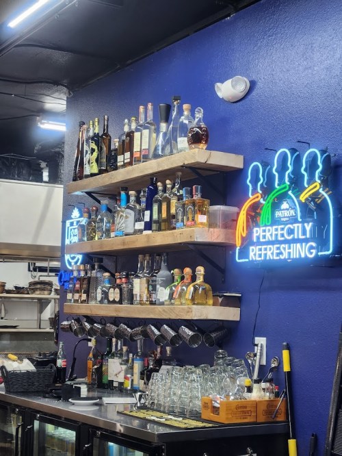A bar shelf with various liquor bottles and a neon sign reading "Perfectly Refreshing" against a blue wall.