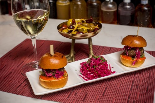 Two mini burgers with pink slaw, a side of vibrant pickled vegetables, and a glass of white wine on a table.