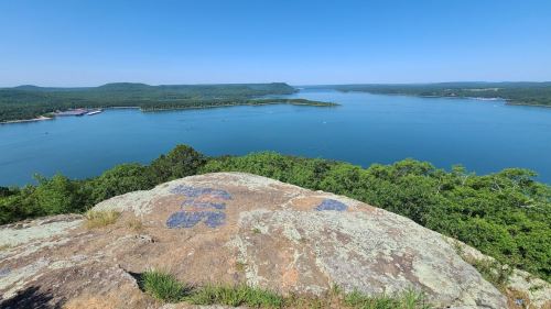 A panoramic view of a serene lake surrounded by lush greenery and distant hills under a clear blue sky.