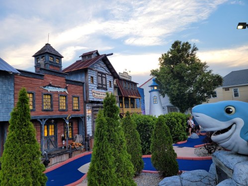 A whimsical mini-golf course featuring colorful buildings, greenery, and a large shark sculpture under a sunset sky.