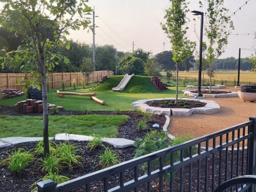 A playground with a grassy hill, slide, logs, and landscaped areas, surrounded by trees and pathways.
