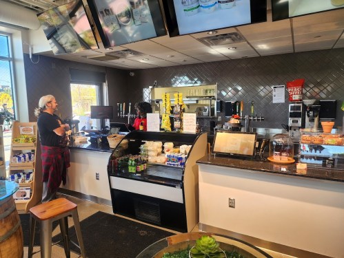 A café interior with a counter, display case, and customers ordering drinks. Screens show menu items above.