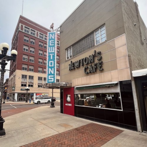 Exterior of Newton's Cafe with a vintage sign, located on a city street with nearby buildings and parked cars.