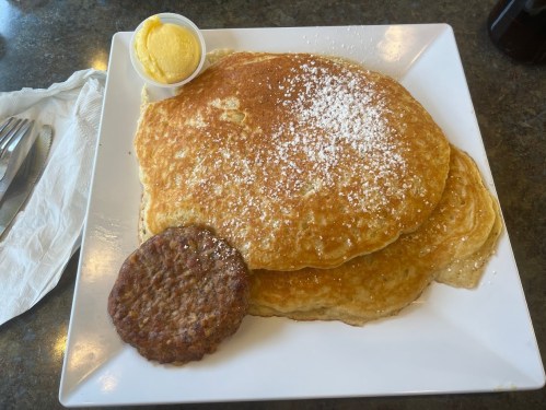 A plate with two large pancakes dusted with powdered sugar, a sausage patty, and a small cup of butter.