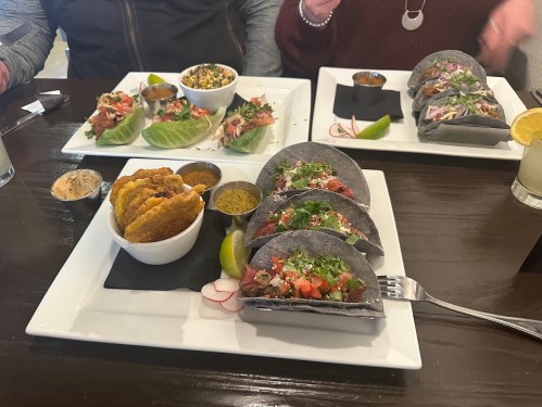 A table with two plates of tacos, a bowl of salad, and a side of fried plantains, garnished with lime and radish.