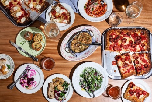 A variety of Italian dishes on a wooden table, including pasta, pizza, salads, and desserts, with glasses of wine.