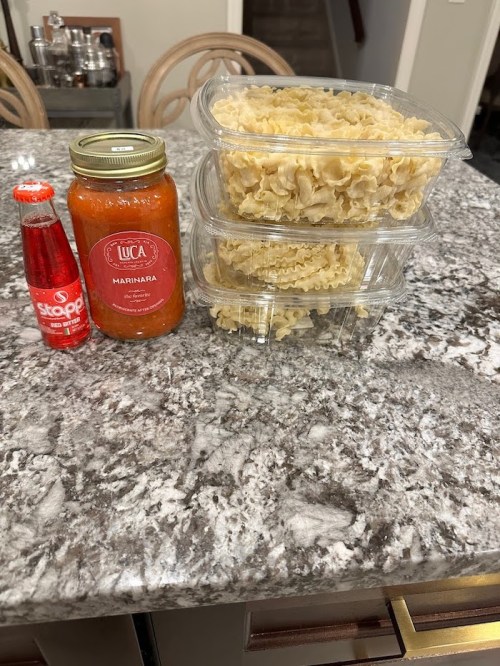 A granite countertop with three containers of cooked pasta, a jar of marinara sauce, and a bottle of soda.