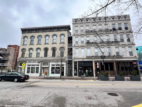 Two historic buildings side by side on a city street, featuring large windows and decorative facades, under a cloudy sky.