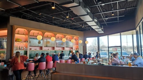 A modern restaurant interior with a bar, pink stools, and patrons enjoying their meals in a bright, spacious setting.