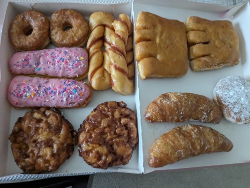 A box of assorted donuts, including glazed, frosted, and filled varieties, alongside croissants and apple fritters.