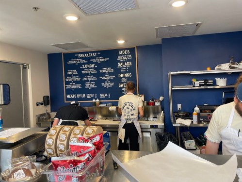 A restaurant kitchen with staff preparing food, a menu board on the wall, and various ingredients on the counter.