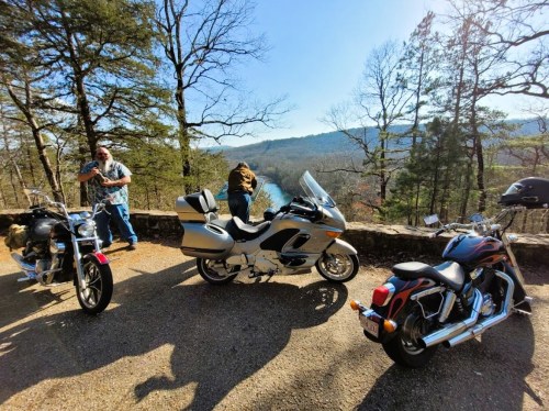 Two motorcycles parked by a scenic overlook, with a man standing nearby and trees in the background.