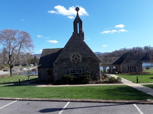 A stone building with a steeple, surrounded by green grass and trees, near a lake under a clear blue sky.