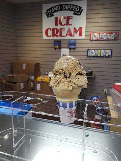 A tall ice cream cone with a scoop of brown ice cream, set against a shop backdrop with an "Ice Cream" sign.