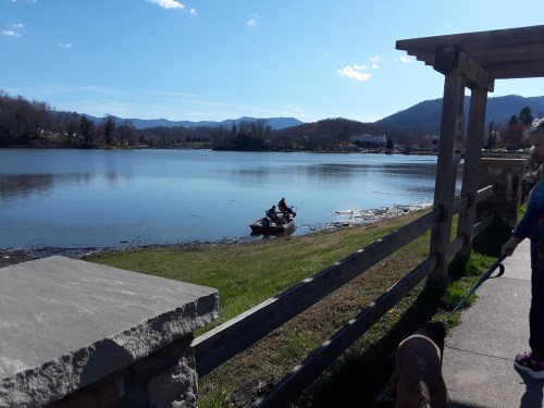 A serene lake scene with mountains in the background, a person in a kayak, and a walking path beside the water.