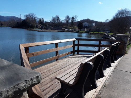 A wooden bench overlooks a calm lake with trees and houses in the background under a clear blue sky.