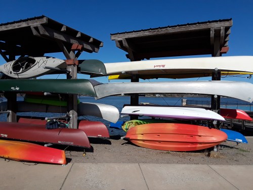 Colorful kayaks and canoes stacked on racks by a calm lake under a clear blue sky.